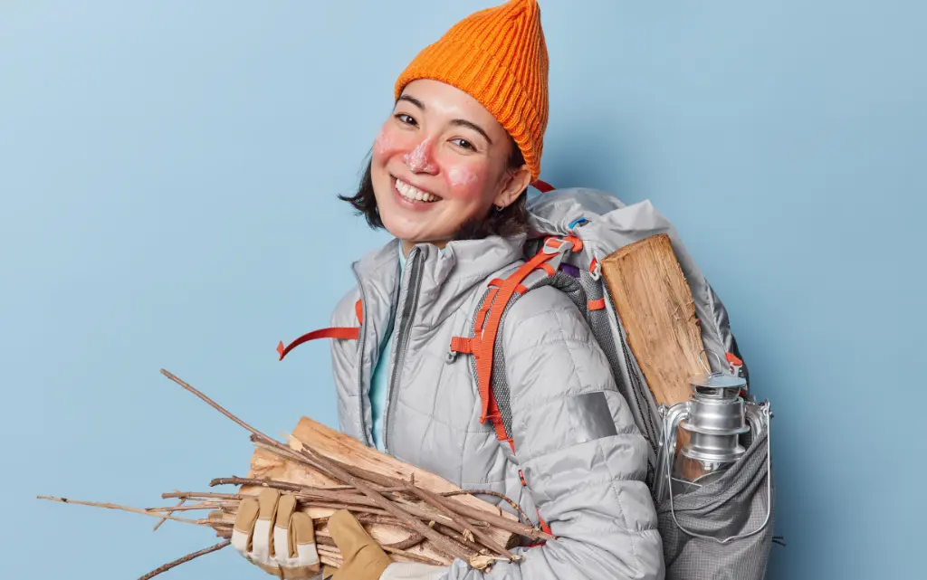 Uśmiechnięta, ubrana w zimowe ubrania kobieta z czerwonymi policzkami, z plecakiem na plecach i chrustem do opału w dłoniach