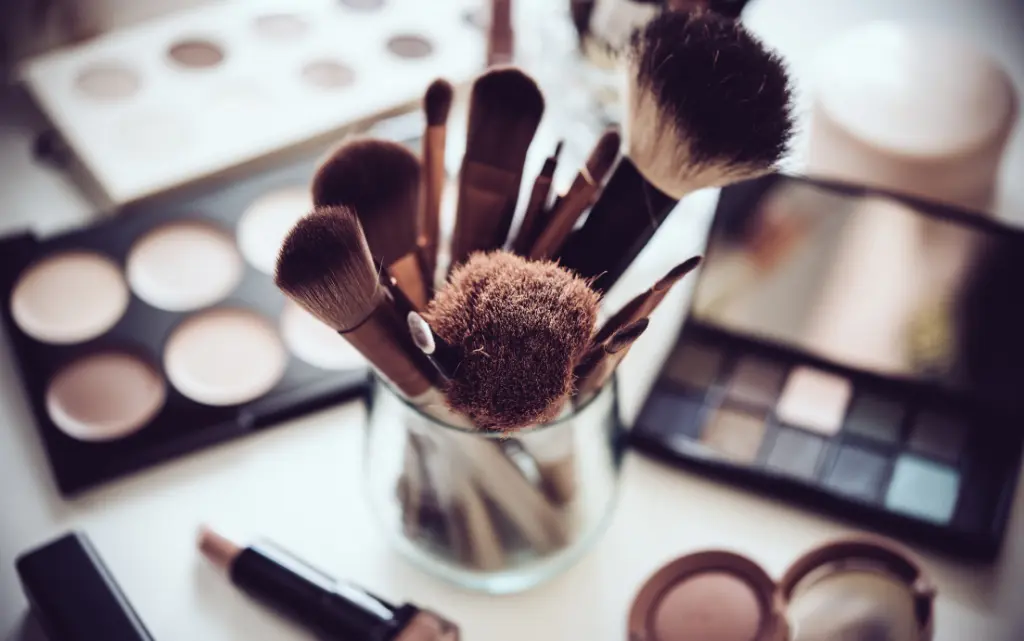 Pędzle do makijażu stojące w kubku, w tle kosmetyki do malowania