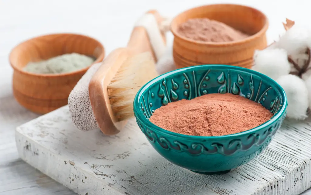 Sproszkowana, czerwona glinka w kamionkowym naczyniu, w tle inne glinki kosmetyczne