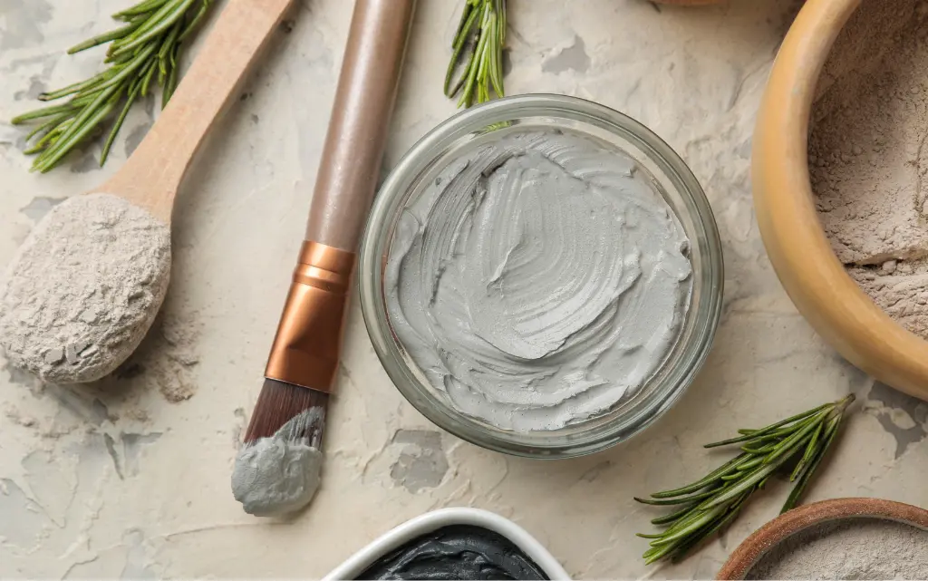 Maseczka z szarej glinki, obok sproszkowana glinka i pędzel do aplikacji maski