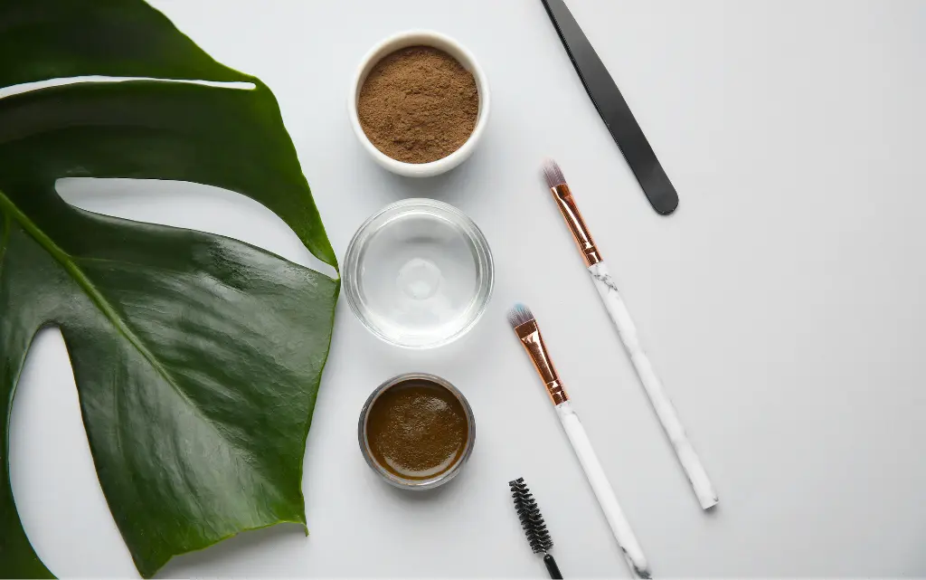 Białe tło, na nim kosmetyki do stylizacji brwi: pudry, woski, cienie, pędzle oraz pęsetka
