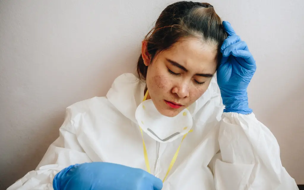 Zmęczona kobieta w białym kombinezonie, rękawiczkach ochronnych zdejmuje maskę ochronną i pokazuje podrażnioną cerę