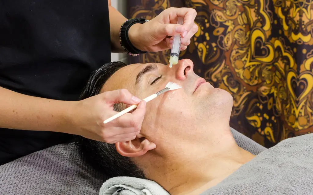 Mężczyzna leży u kosmetyki i poddaje się zabiegowi oczyszczania twarzy, kobieta nakłada pędzelkiem kwas glikolowy