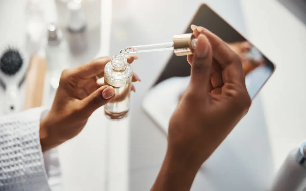 Kobiece dłonie trzymające serum z pipetką, w tle lusterko kosmetyczne i szczotka do włosów