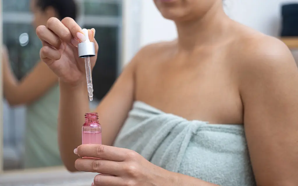 Kobieta w ręczniku w łazience trzyma w dłoni różową butelkę, a w drugiej dłoni pipetkę kosmetyczną