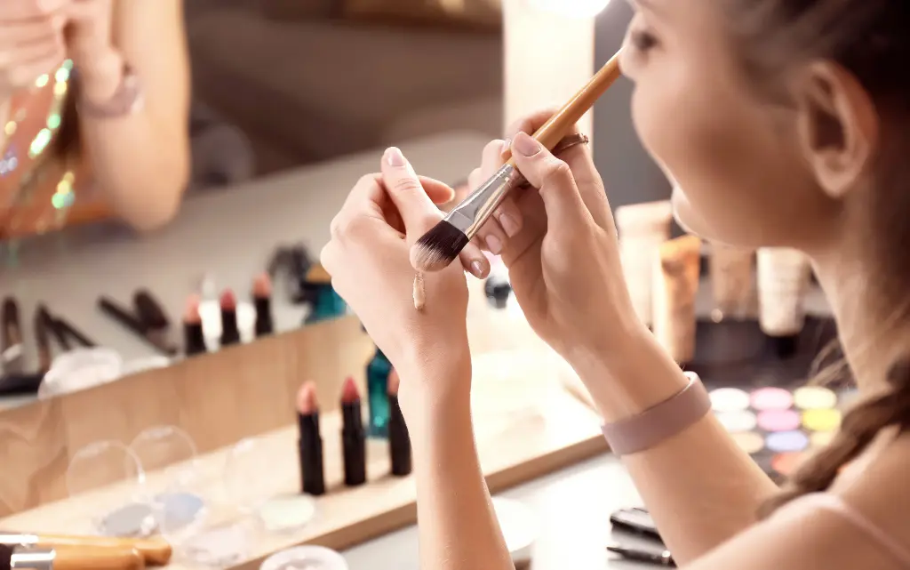 Dziewczyna przed toaletką z kosmetykami nakłada na dłoń pędzlem podkład do cery tłustej