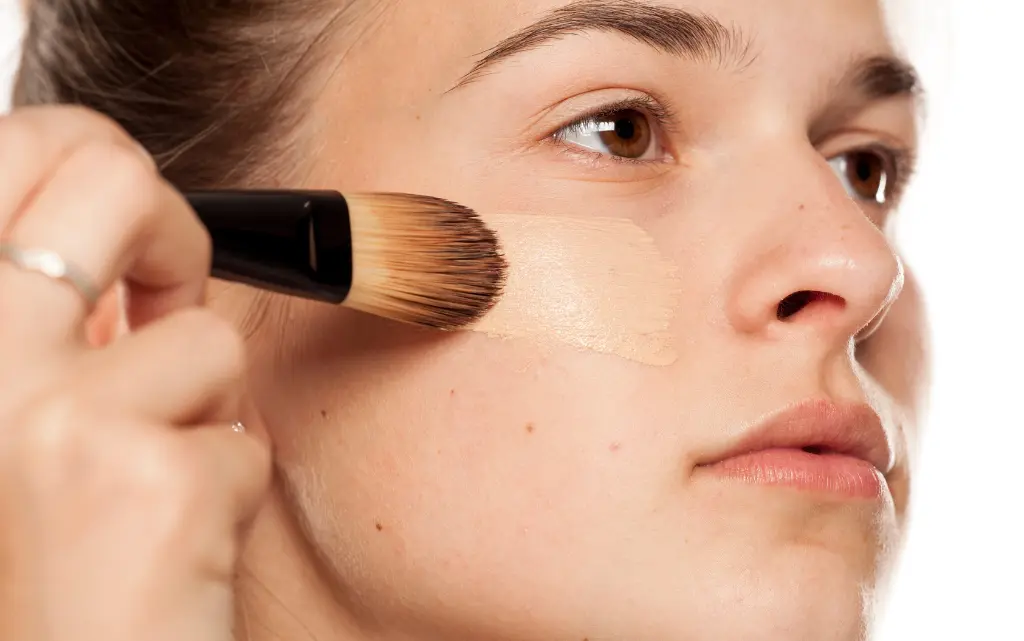Młoda dziewczyna nakłada przy pomocy pędzla do makijażu podkład nawilżający