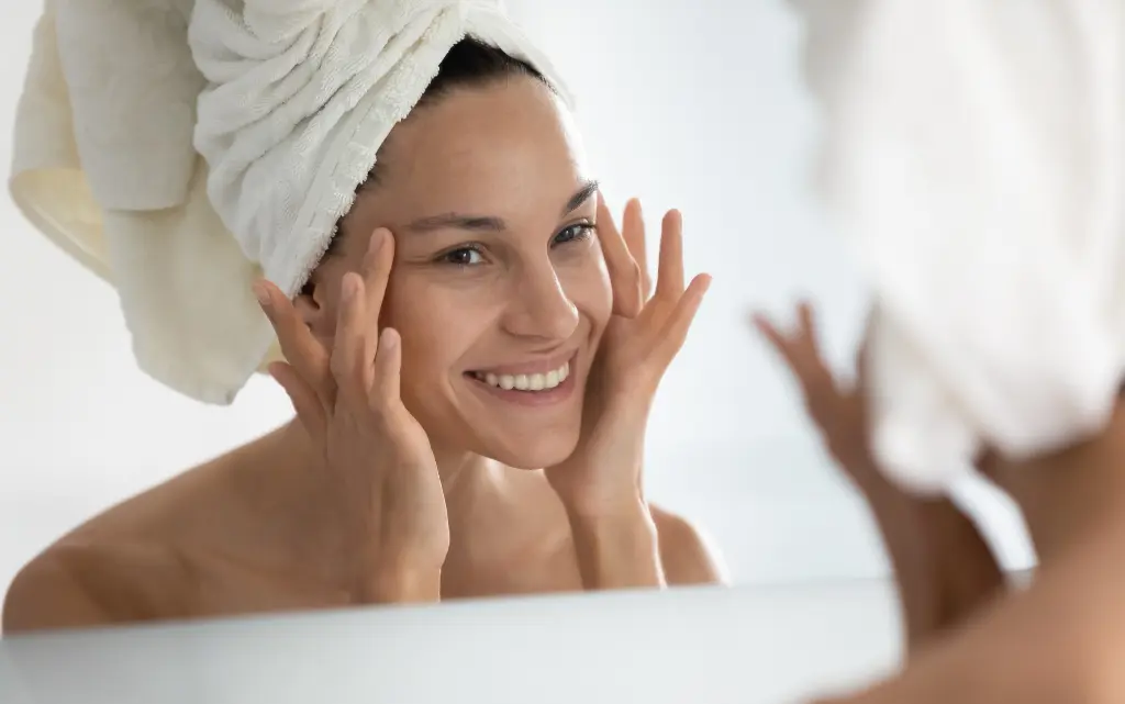 Uśmiechnięta kobieta przed lustrem z włosami w ręczniku dotyka swojej twarzy