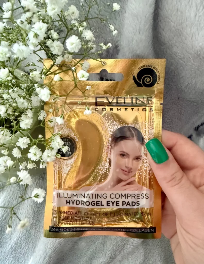 Hydrożelowe płatki pod oczy kompres rozświetlający Eveline trzymane przez kobiecą dłoń na tle białych kwiatów