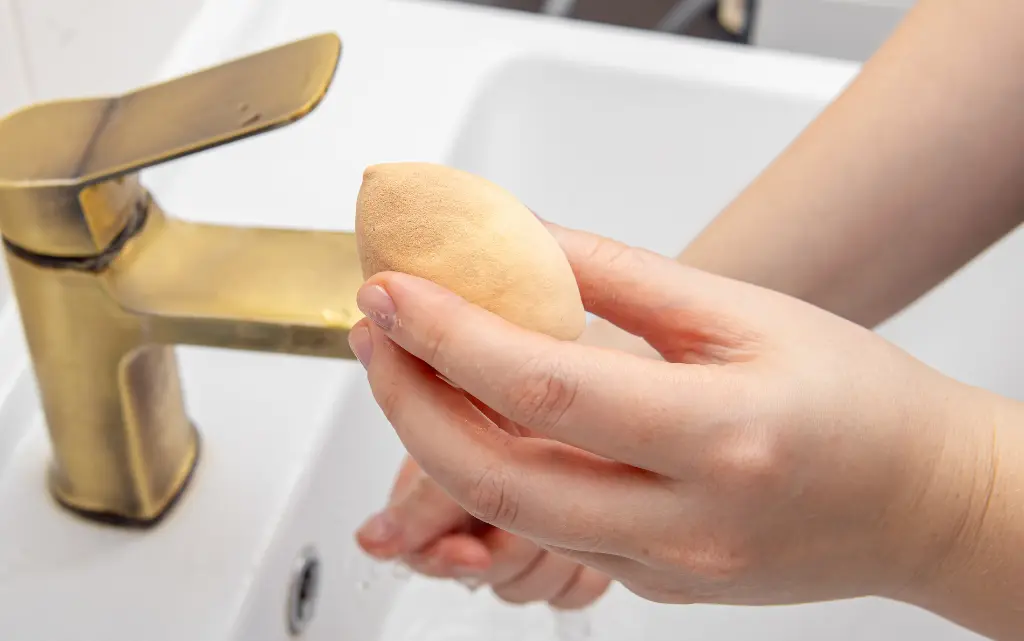 Brudna gąbka do makijażu w rękach kobiety, w tle złota bateria umywalkowa
