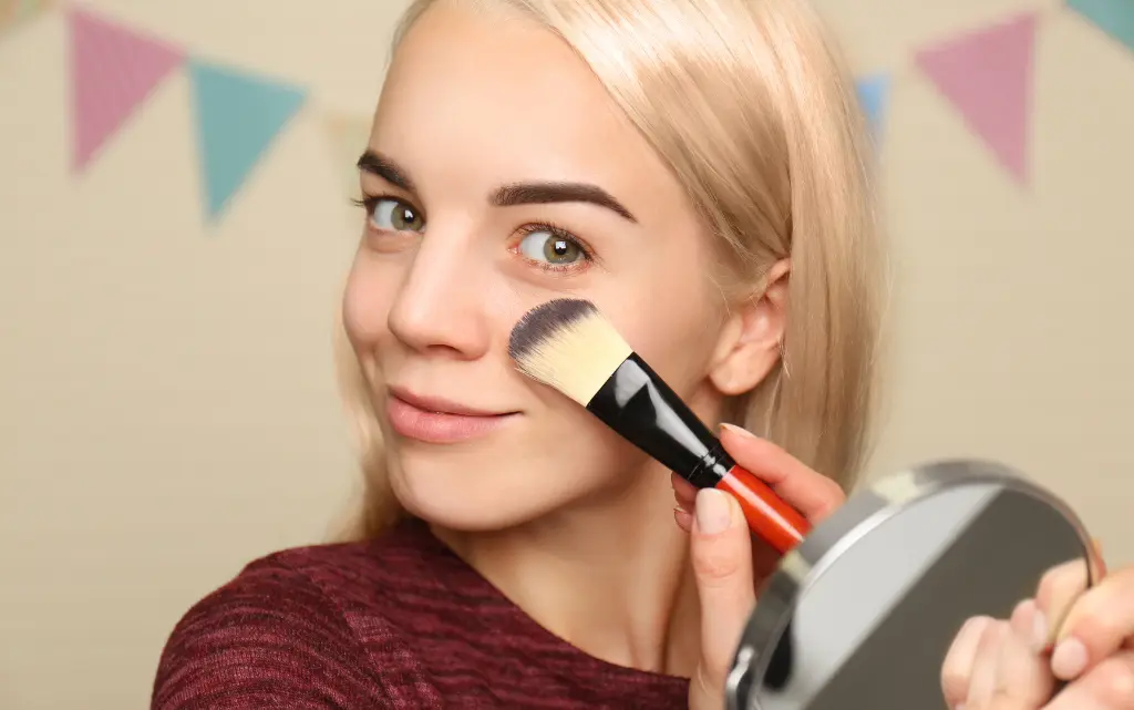 Młoda dziewczyna trzyma pędzel do makijaży i uśmiecha się do lusterka