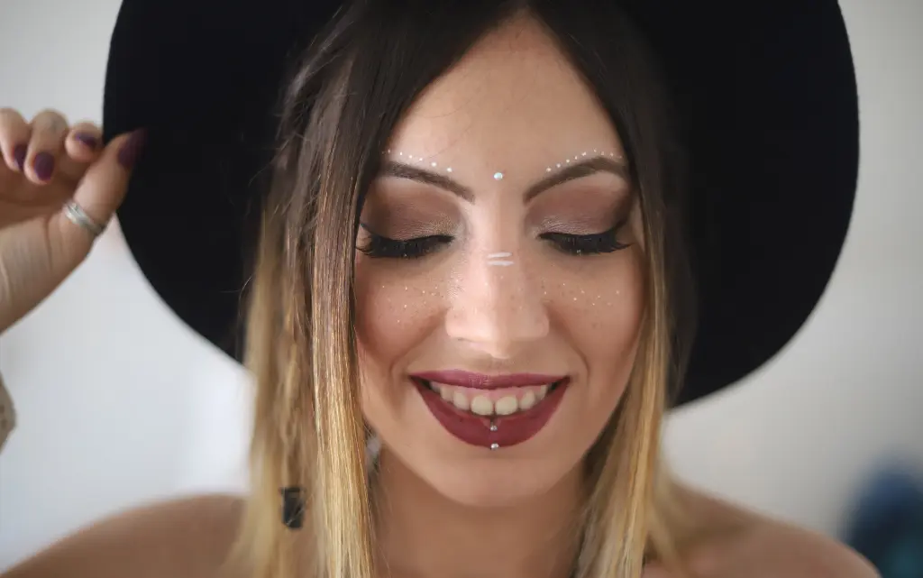 Kobieta w czarnym kapeluszu w makijażu festiwalowym z mocno zaznaczonymi ustami i diamencikami wzdłuż linii brwi