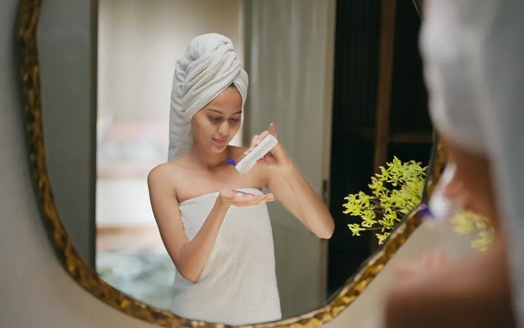 Kobieta po kąpieli przed lustrem nakłada na dłoń kremowy tonik do cery suchej