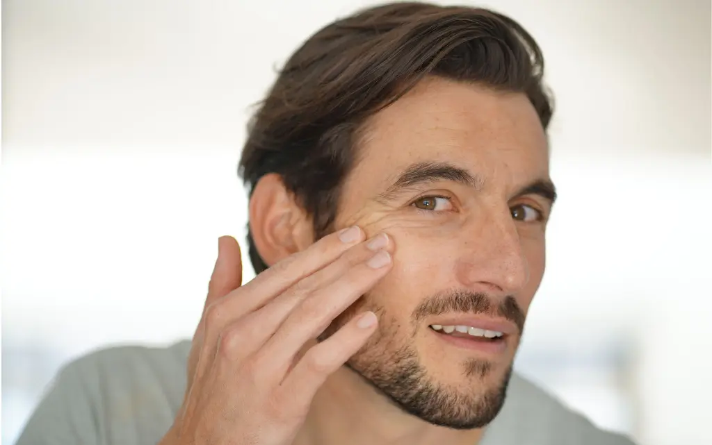 Mężczyzna z zarostem przed lustrem analizuje swoją skórę twarzy i zmarszczki