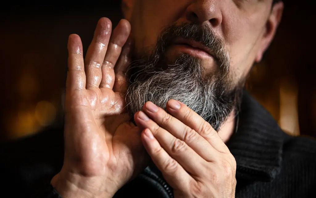 Mężczyzna aplikuje na brodę olejek, ma tłuste dłonie od kosmetyku do brody