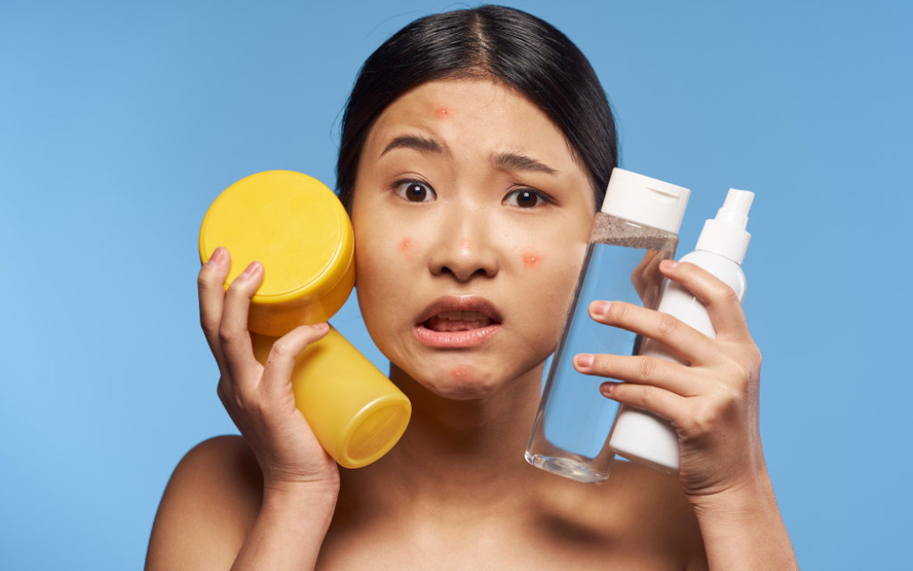 Zagubiona dorosła kobieta z trądzikiem dorosłych trzyma przy twarzy różne opakowania kosmetyków do cery trądzikowej