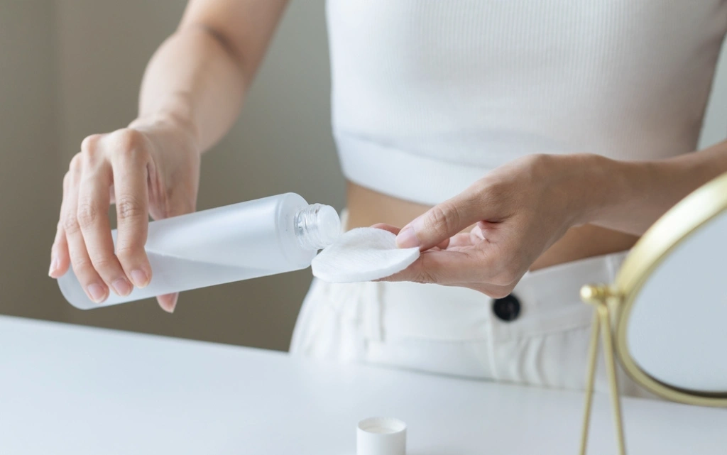 Młoda kobieta w białym ubraniu nalewa na wacik tonik oczyszczający do twarzy