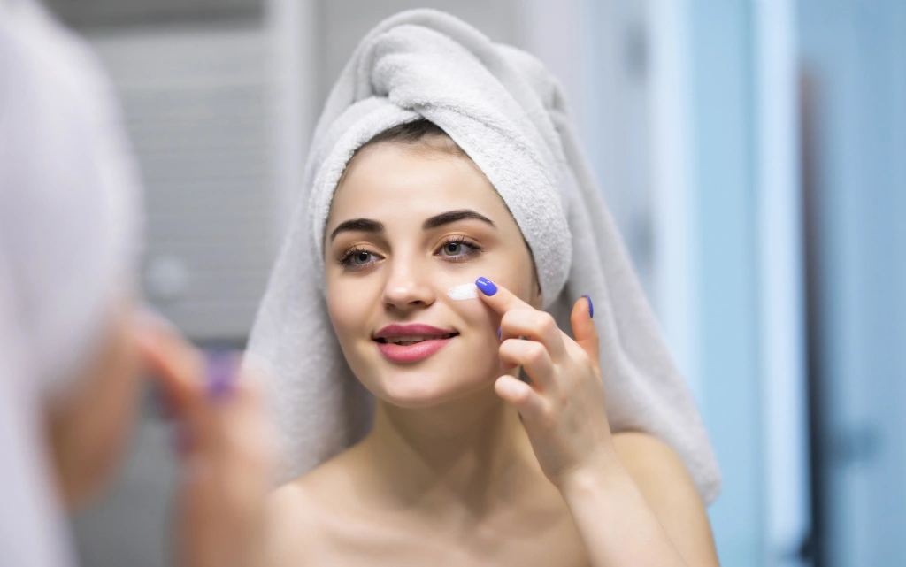 Młoda 0-letni kobieta w ręczniku na włosach nakłada krem przeciwzmarszczkowy na twarz