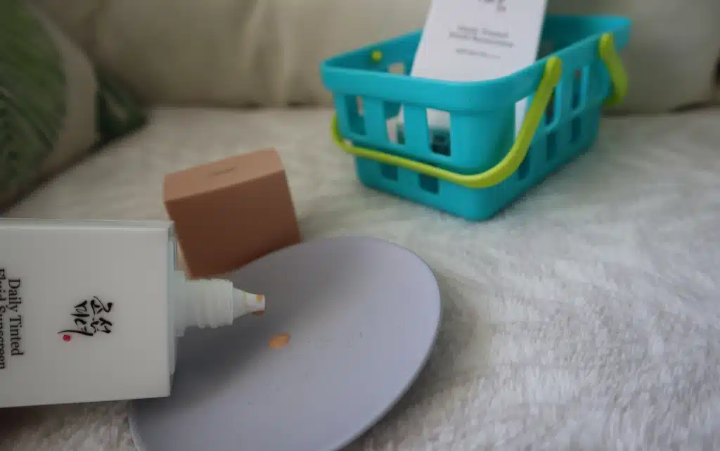 Otwarta buteleczka kremu na słońce Daily Tinted Fluid Sunscreen Beauty of Joseon pokazująca lejącą konsystencję produktu, w tle niebieski koszyczek i poduszki