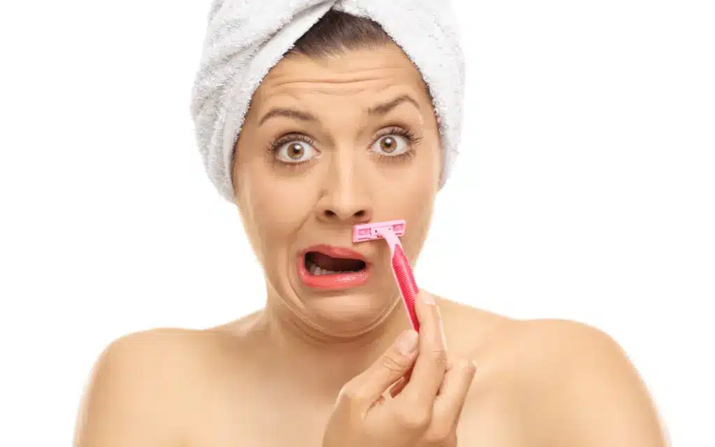 Kobieta z wyraźnie niezadowoloną miną usuwa włoski na twarzy z pomocą maszynki jednorazowej do golenia