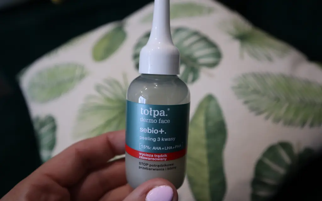 Dłoń trzymająca Tołpa Sebio+ Peeling 3 Kwasy na tle beżowo-zielonej poduszki