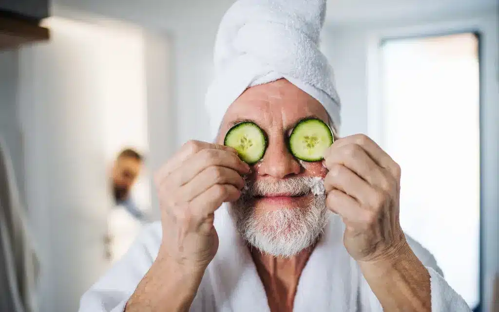 Starszy mężczyzna z białą brodą, ręcznikiem na głowie trzymający plasterki ogórka na oczach