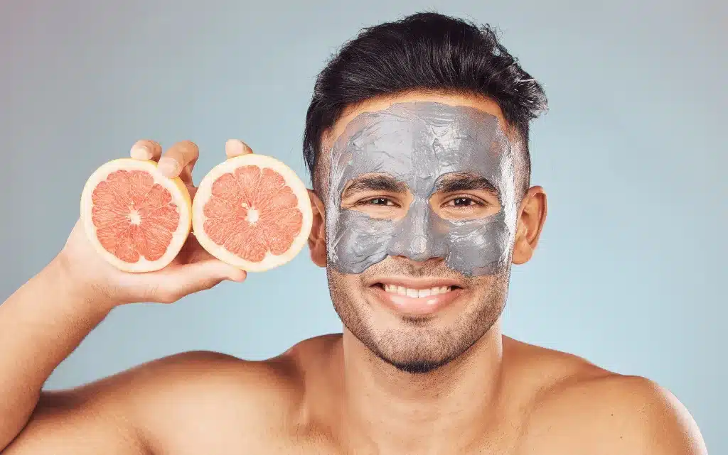 Uśmiechnięty mężczyzna z peelingiem na twarzy, trzyma w dłoniach przekrojony grejpfrut
