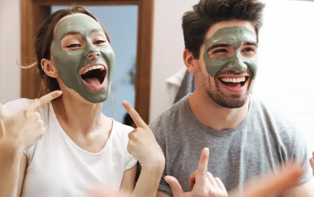 Mężczyzna i kobieta w maseczce z zielonej glinki uśmiechają się przed lustrem w łazience