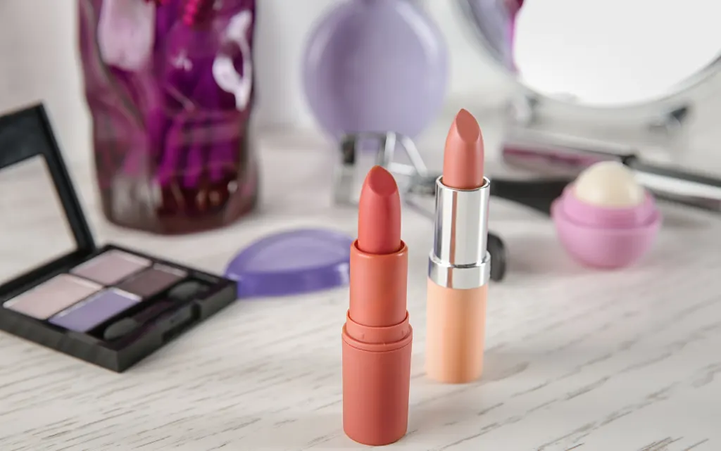 Dwie kolorowe szminki do ust, w tle cienie do powiek, balsam do ust i akcesoria do makijażu