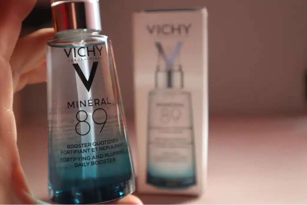 Kobieca dłoń trzyma szklaną butelkę Vichy Minéral 89 - Booster wzmacniająco-nawilżający, w tle papierowe opakowanie
