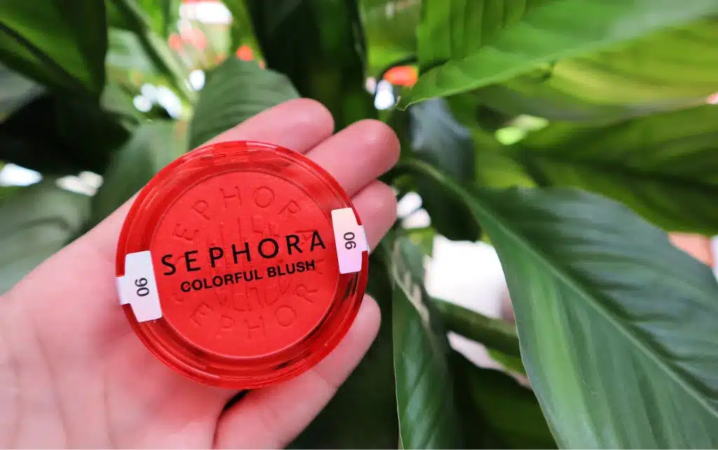 W kobiecej dłoni Rozświetlający róż w pudrze Sephora Collection Colorful Blush 06 Tropical Dawn na tle zielonych liści