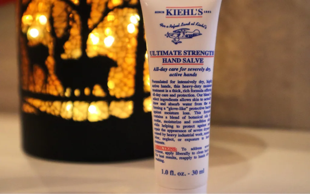 Krem do rąk Kiehl's Ultimate Strenght Hand Salve na tle świecącej, dekoracyjnej lampki