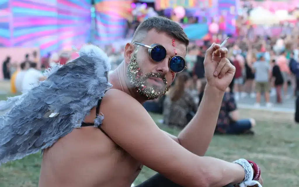 Męzczyzna ze skrzydłami anioła pali papierosa na festiwalu, ma czarne okulary i makijaż z diamencikami na twarzy