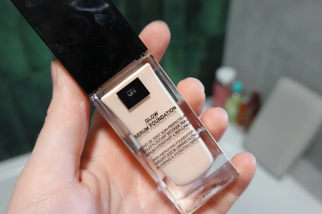 Podkład poprawiający wygląd skóry Givenchy Prisme Libre Glow Serum w kobiecej dłoni, w tle łazienka