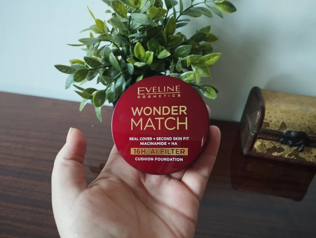 Eveline Cosmetics Wonder Match Kryjący podkład cushion trzymany w kobiecej dłoni, w tle zielony kwiatek