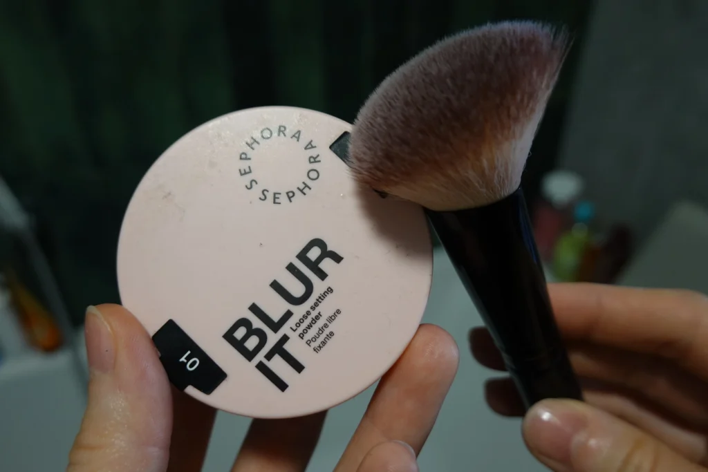 Sephora Collection Blur It Sypki Puder Utrwalający trzymany w kobiecej dłoni, obok pędzel do pudru