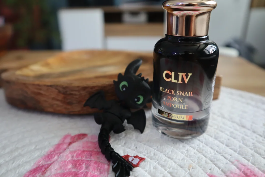 Buteleczka CLIV Black Snail PDRN Ampoule ze złotą zakrętką stojąca na drewnianej desce obok czarnej figurki smoka Szczerbatka