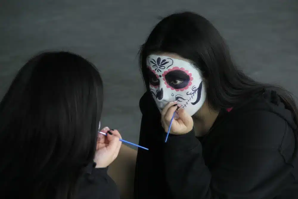 Wizażystka maluje cienkim pędzlem makijaż kościotrupa w stylu sugar skull — różowe łuski wokół oczu i ornament na czole 